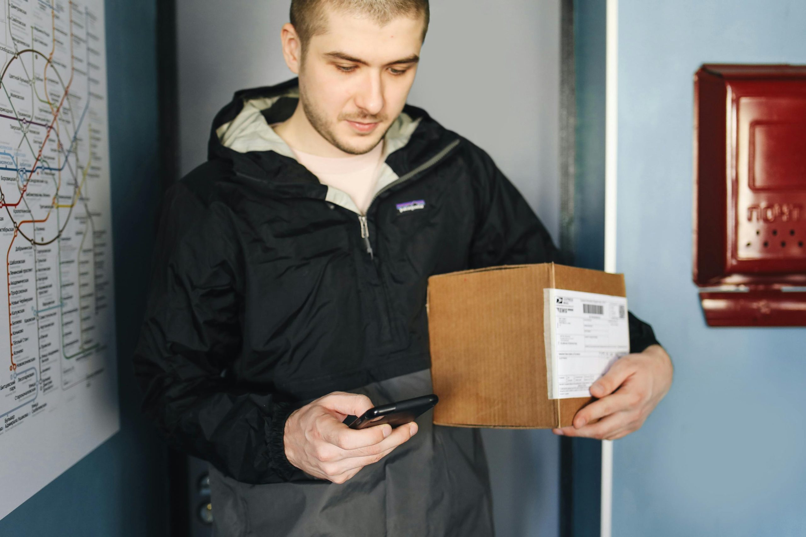 Man in casual jacket scans package label using smartphone indoors near metro map.