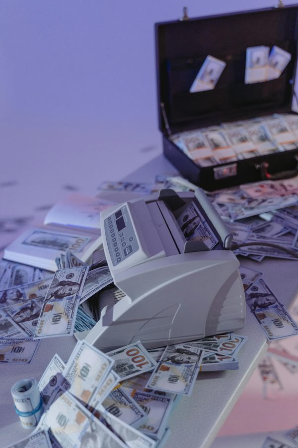 A money counter surrounded by scattered dollar bills on a table with a suitcase full of cash.